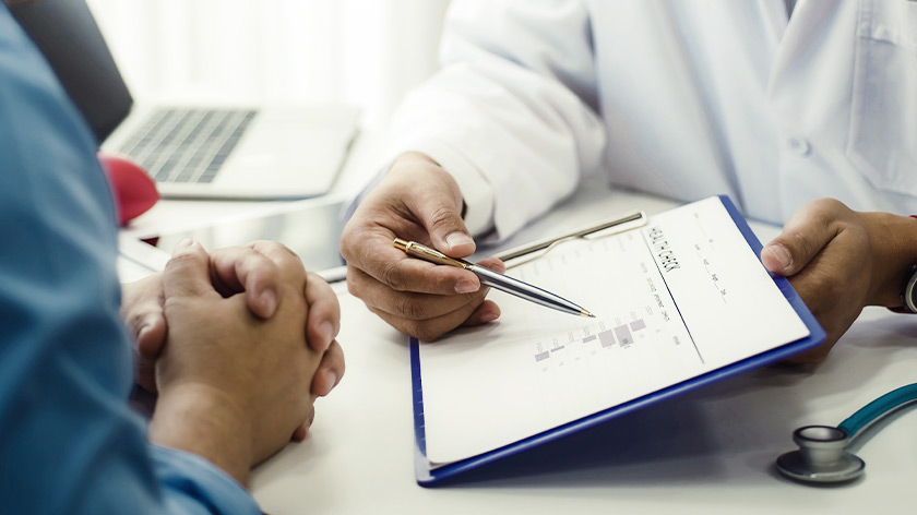 Arzt sitzt Patienten am Tisch gegenüber und zeigt mit Stift auf ein Tablet, das er in der Hand hält